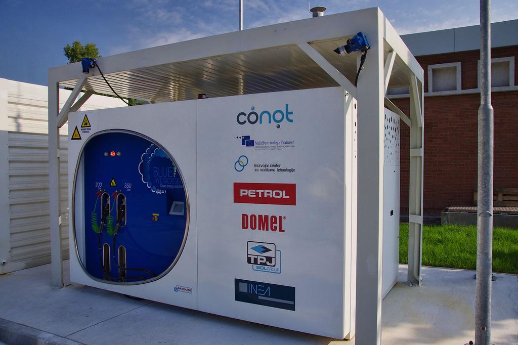 Na fotografiji je prva vodikova polnilnica v Sloveniji, postavljena na Petrolovem bencinskem servisu v Lescah. Na sliki je bela modularna enota z modro okroglo nadzorno ploščo in logotipi, med katerimi so Conot, Petrol in Domel. Ob njej stoji velik bel zaboj z varnostnimi opozorili. Celoten sistem je umeščen na urejeni tlakovanji površini z označenimi parkirnimi mesti, v ozadju pa so vidne stavbe, ograja in zelenje pod delno oblačnim nebom.