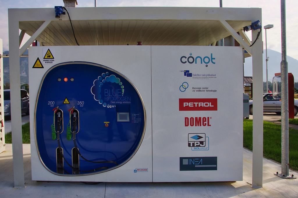 Na fotografiji je prva vodikova polnilnica v Sloveniji, postavljena na Petrolovem bencinskem servisu v Lescah. Na sliki je bela modularna enota z modro okroglo nadzorno ploščo in logotipi, med katerimi so Conot, Petrol in Domel. Ob njej stoji velik bel zaboj z varnostnimi opozorili. Celoten sistem je umeščen na urejeni tlakovanji površini z označenimi parkirnimi mesti, v ozadju pa so vidne stavbe, ograja in zelenje pod delno oblačnim nebom.