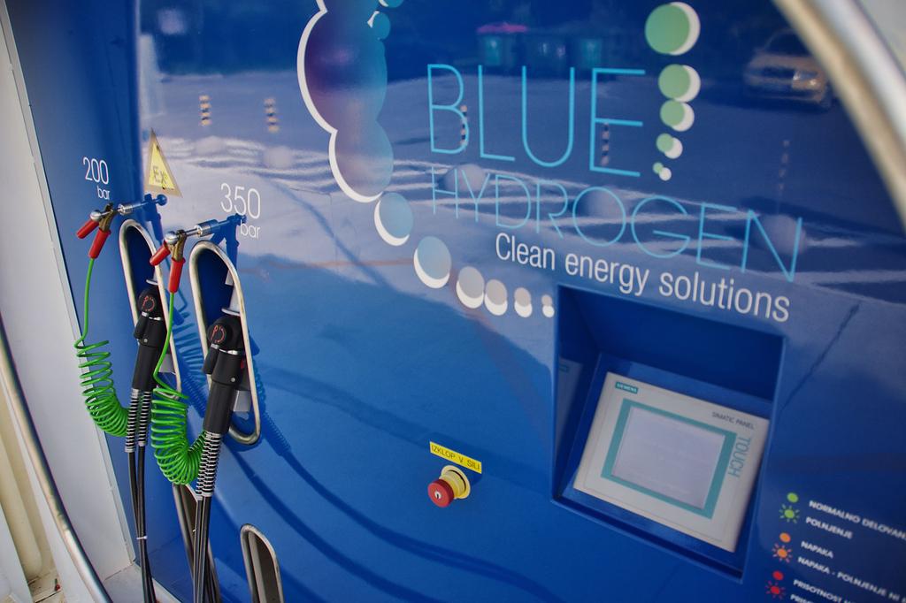 Na fotografiji je bližnji posnetek modre vodikove polnilne enote z napisom 'BLUE HYDROGEN – Clean energy solutions'. Na levi strani sta dve polnilni pištoli z zelenimi spiralnimi kabli, označeni z vrednostma 200 bar in 350 bar. Spodaj je nameščen rdeč varnostni gumb, desno pa digitalni upravljalni zaslon. Gre za del prve slovenske vodikove polnilnice na Petrolovem servisu v Lescah.