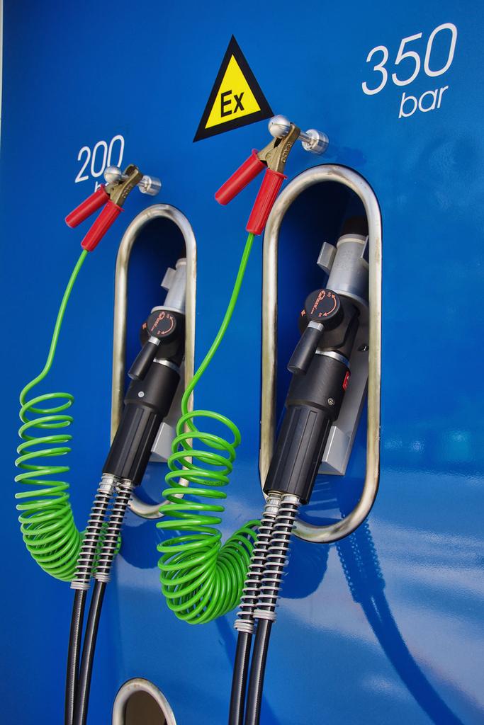 Na fotografiji je bližnji posnetek modre vodikove polnilne enote z napisom 'BLUE HYDROGEN – Clean energy solutions'. Na levi strani sta dve polnilni pištoli z zelenimi spiralnimi kabli, označeni z vrednostma 200 bar in 350 bar. Spodaj je nameščen rdeč varnostni gumb, desno pa digitalni upravljalni zaslon. Gre za del prve slovenske vodikove polnilnice na Petrolovem servisu v Lescah.