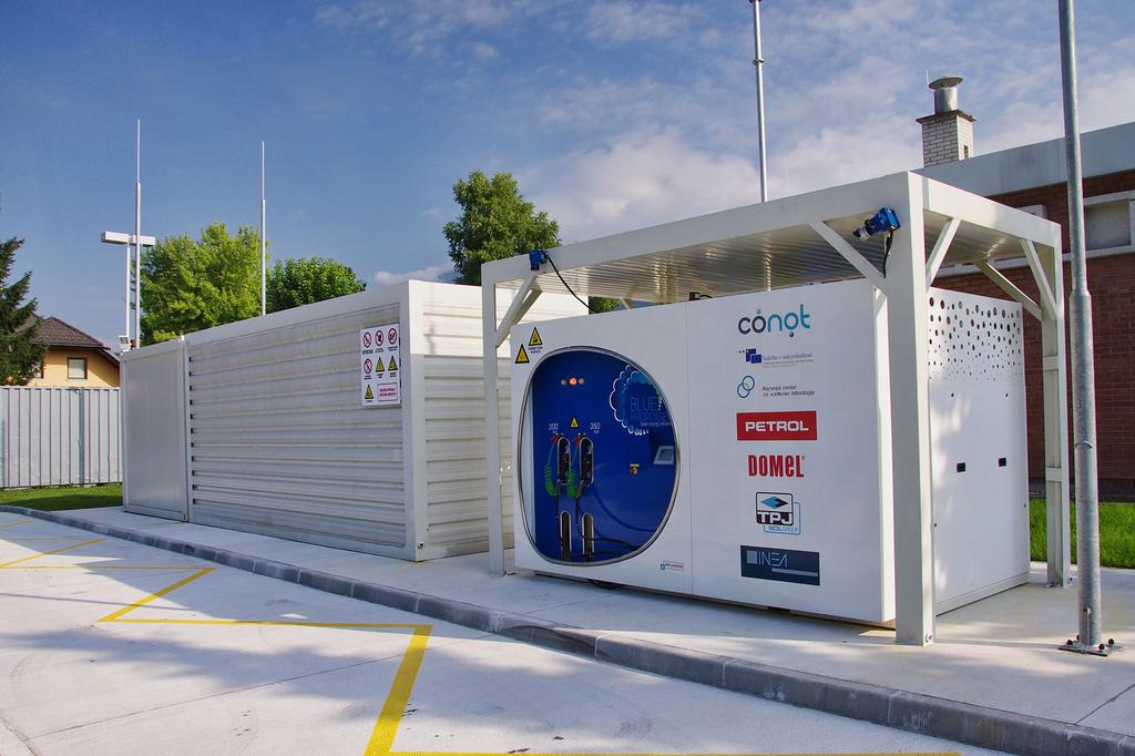 Na fotografiji je prva vodikova polnilnica v Sloveniji, postavljena na Petrolovem bencinskem servisu v Lescah. Na sliki je bela modularna enota z modro okroglo nadzorno ploščo in logotipi, med katerimi so Conot, Petrol in Domel. Ob njej stoji velik bel zaboj z varnostnimi opozorili. Celoten sistem je umeščen na urejeni tlakovanji površini z označenimi parkirnimi mesti, v ozadju pa so vidne stavbe, ograja in zelenje pod delno oblačnim nebom.