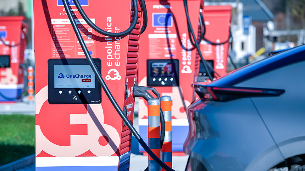 Na sliki so rdeče električne polnilnice za vozila z zasloni ‘OneCharge’ in kabli, ob katerih je parkiran električni avtomobil med polnjenjem